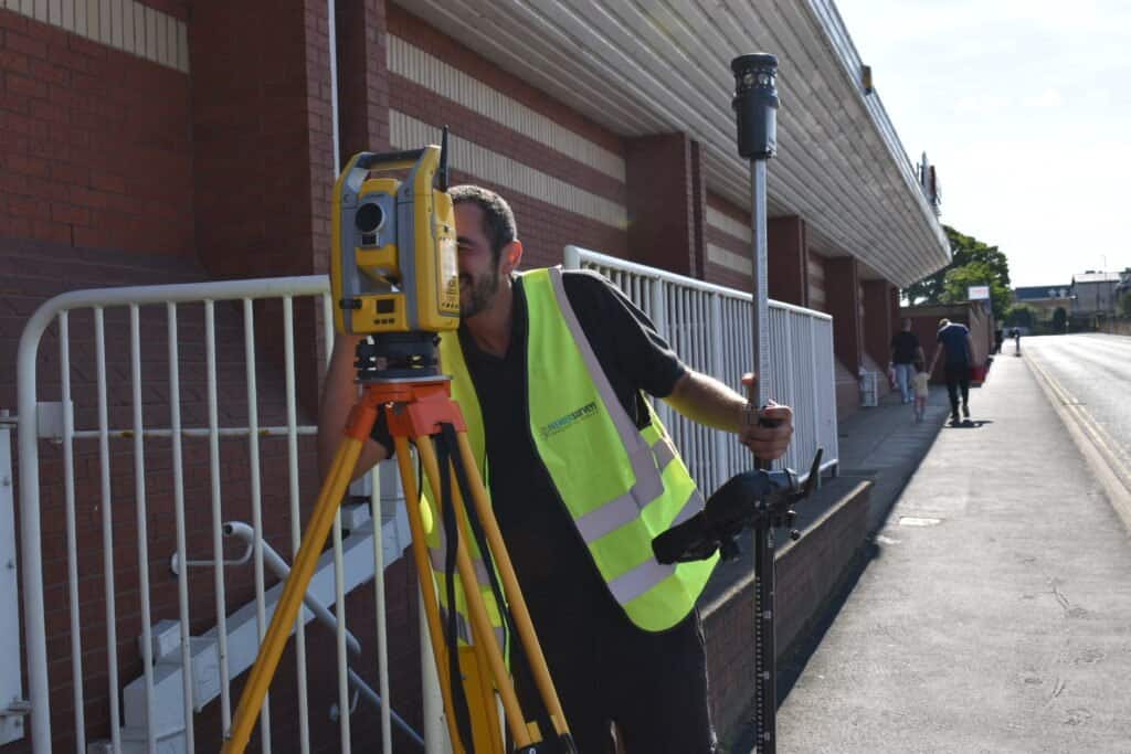 Surveyor using a total station for topographical survey on a city sidewalk with a building in the background.