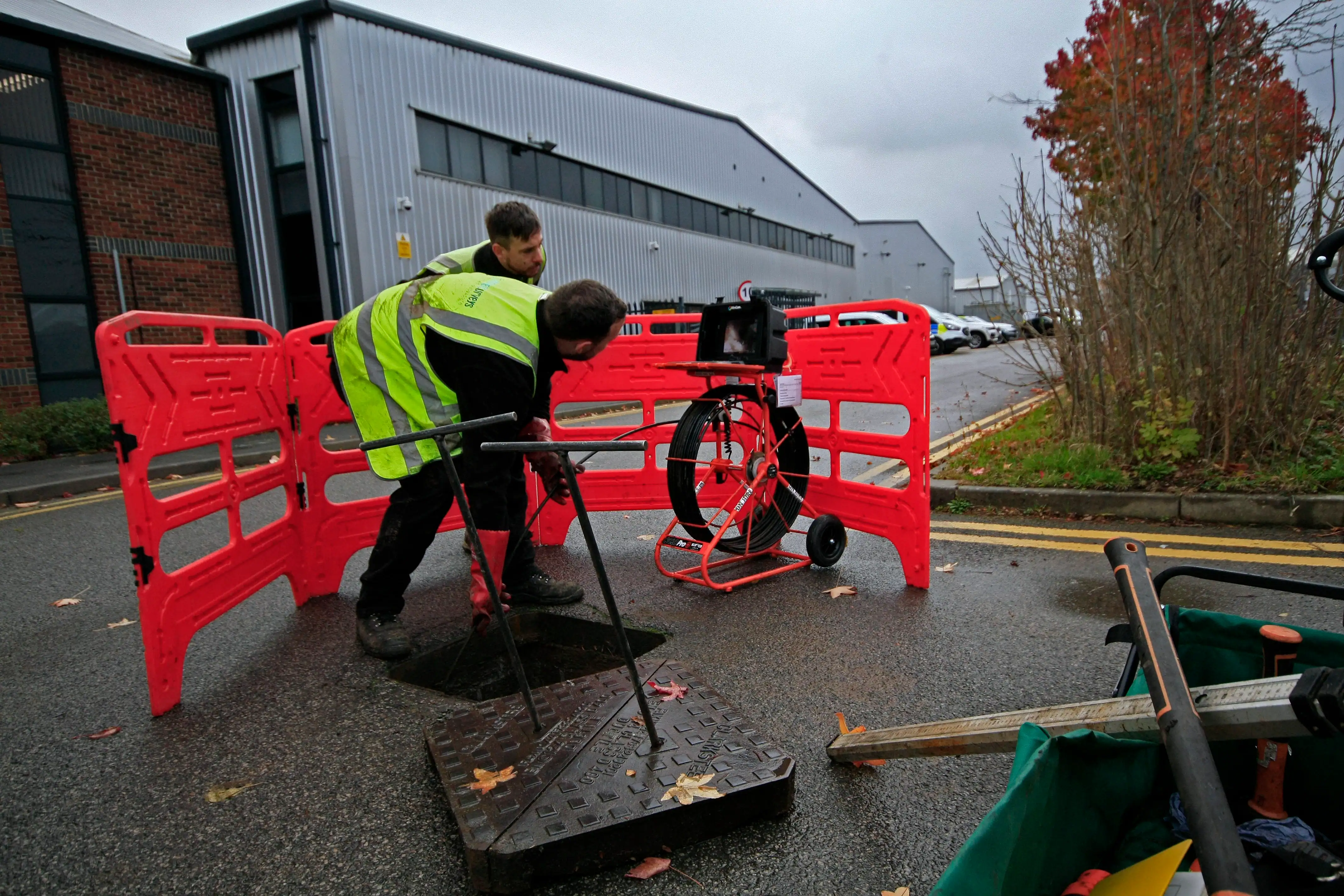 Technicians conducting a CCTV drainage survey with specialized camera equipment behind safety barriers on a cloudy day.