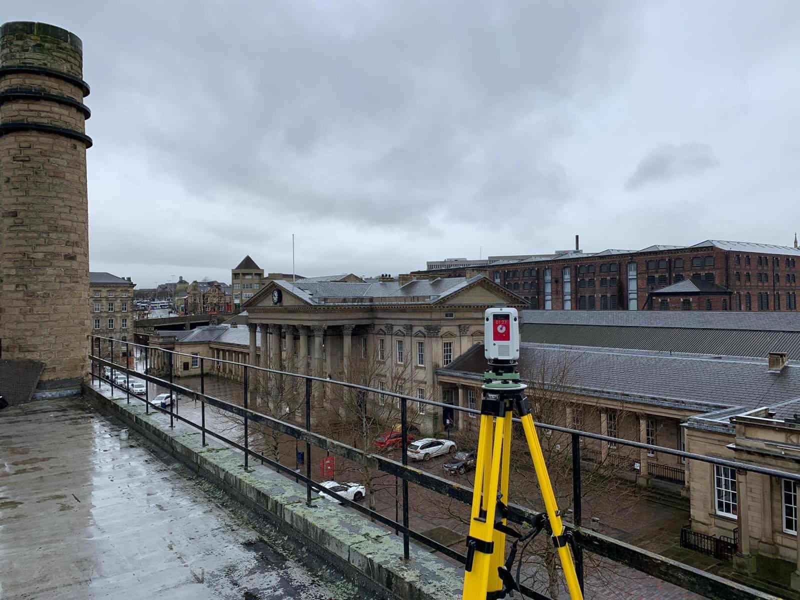 A surveying instrument mounted on a tripod on a rooftop, overlooking historic buildings in an urban area under cloudy skies, used for precise measurements in a measured building survey.